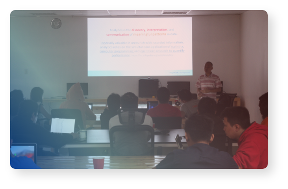 Seated people in the foreground, a person presenting in the background.